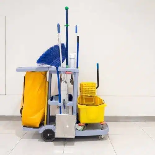 A janitor cart with cleaning supplies, including a mop, broom, yellow bucket, and yellow trash bag, stands against a white wall on a tiled floor—ready for office cleaning or janitorial service tasks.