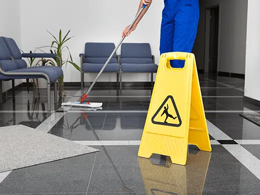 A janitorial service worker mops a shiny floor in a waiting area with blue chairs, while a yellow caution wet floor sign stands prominently in the foreground.