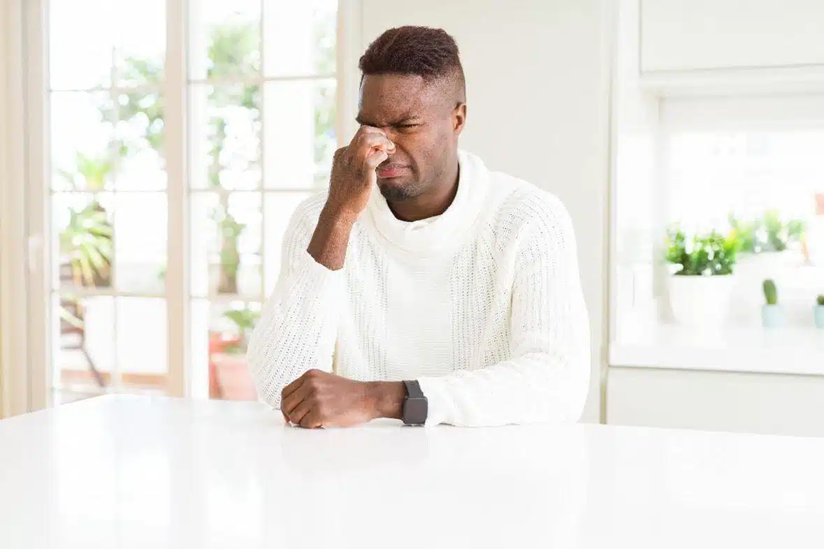 A man in a white sweater sits at a table indoors, pinching the bridge of his nose with his eyes closed, appearing stressed—perhaps after managing commercial cleaning or janitorial service tasks.