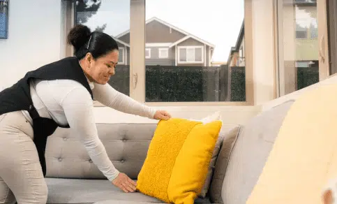 A woman arranges a yellow pillow on a light gray sofa in a living room with large windows and houses visible outside.