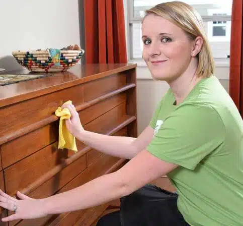 A woman in a green shirt kneels and wipes a wooden dresser with a yellow cloth in a well-lit room.