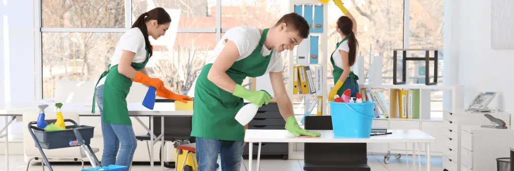 Three people wearing green aprons and gloves are cleaning an office with spray bottles, cloths, and cleaning supplies, showcasing the professionalism of business cleaning services Christiansburg.