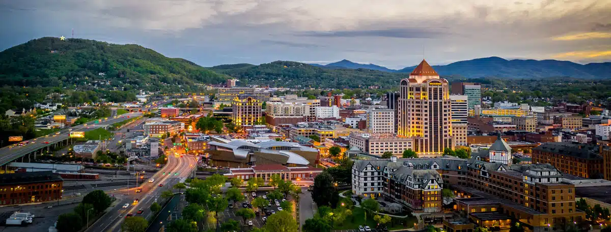 Aerial view of a city with illuminated buildings, roads, and surrounding green hills under a cloudy sky at dusk, showcasing the vibrant atmosphere where Cleaning Service Blacksburg keeps spaces spotless.
