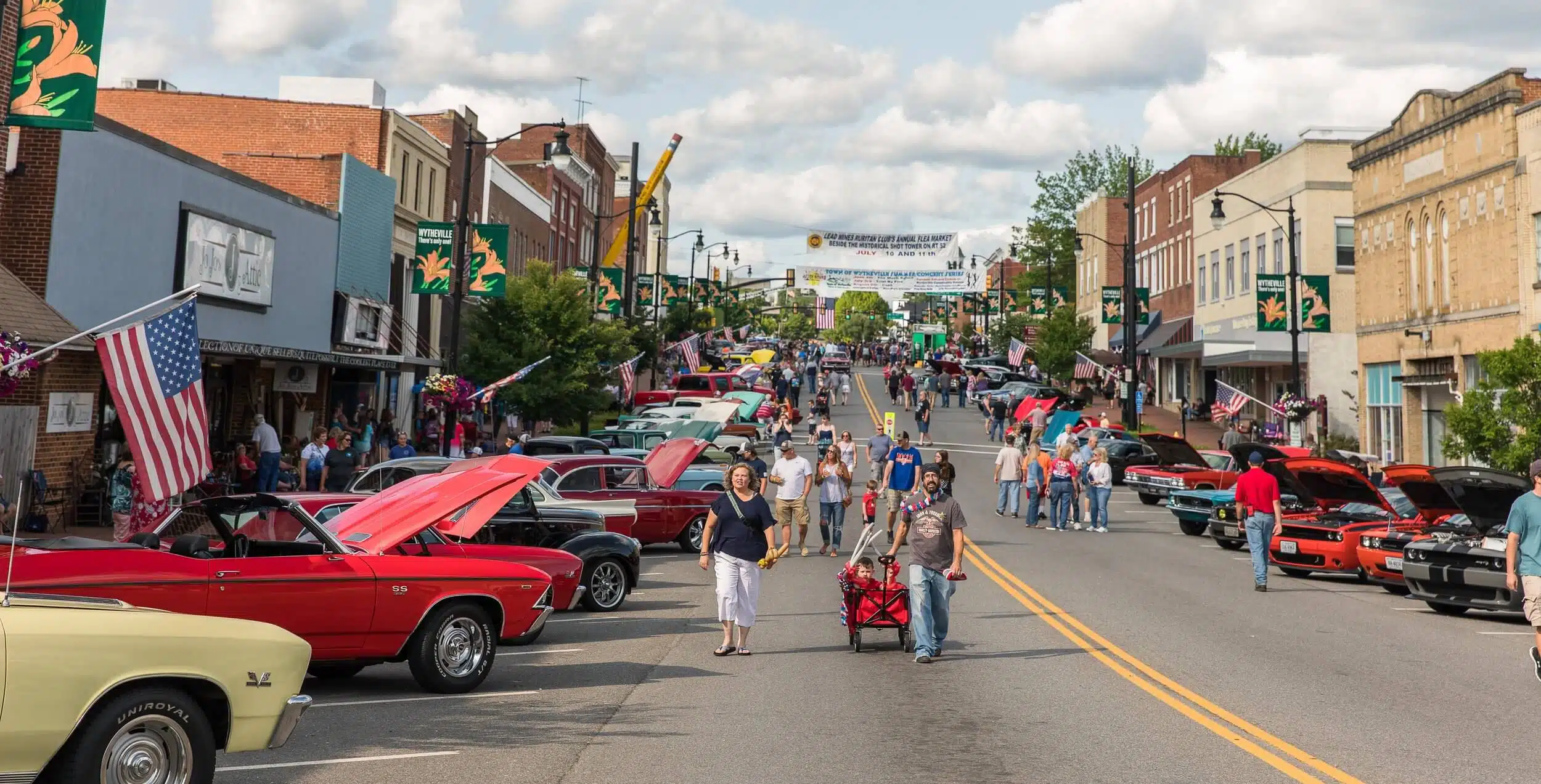 People stroll down a closed-off street lined with classic cars at a small-town car show. Buildings, banners, and American flags are visible, showcasing community pride—just like Cleaning Service Blacksburg brings shine to the heart of town.