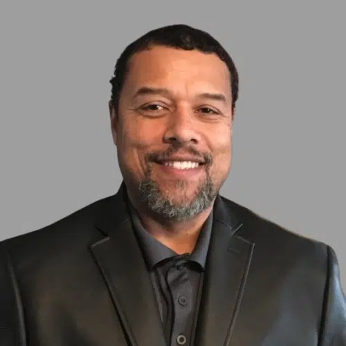 A man with short hair and a beard is smiling, wearing a dark blazer and collared shirt, representing Our Team in front of a plain gray background.